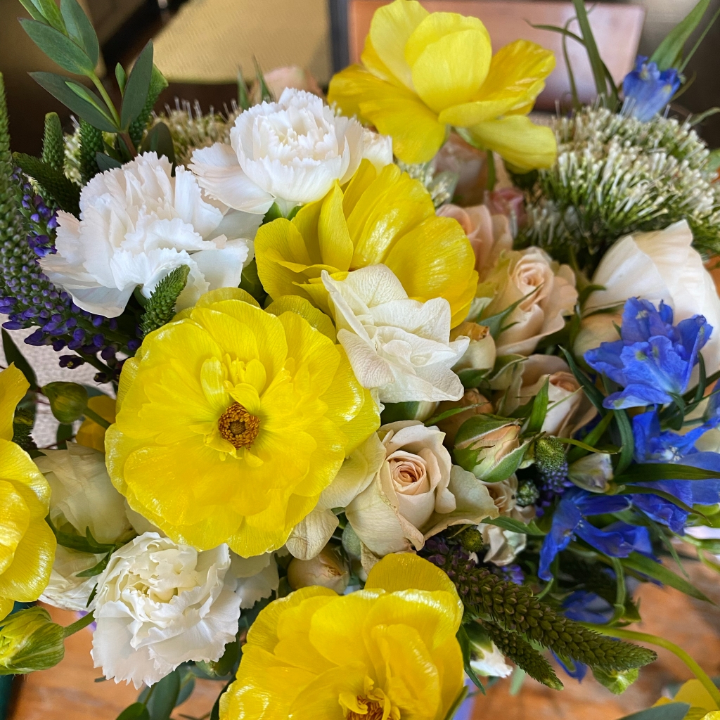 A top-down shot of a bouquet of yellow and white flowers, with some blue and blush pink accents. 