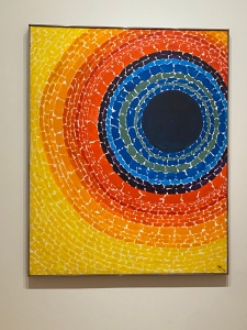 Eclipse, by Alma Thomas. A rectangular canvas features a large, dark blue circle slightly off-center. Radiating out from it are concentric circles made of dashes in shades of blue, orange, and yellow.