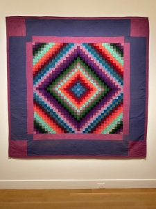 A quilt in the Sunshine and Shadow / A Trip Around the World pattern from an unknown maker in Lancaster County, PA. The square quilt features a mauve binding with squares at each corner, and a lavender surround. The pieced portion of the quilt is made up of tiny squares arranged to form a diamond shape that looks like it's moving. it uses purples, blues, greens, red, coral, and mint.
